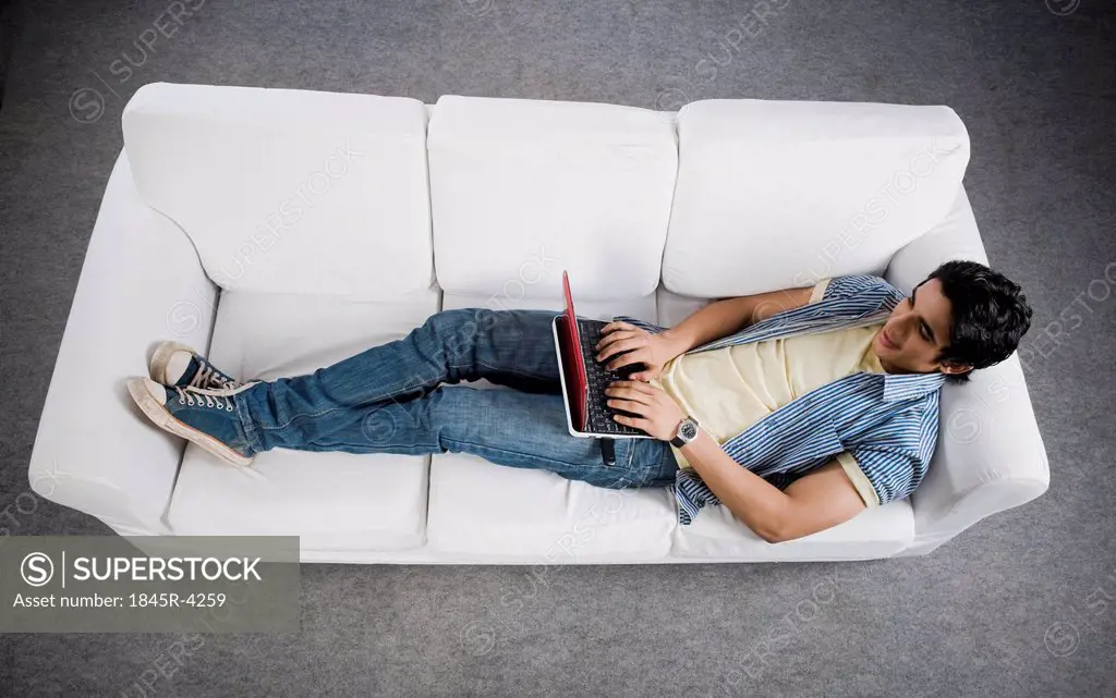 High angle view of a man lying on a couch working on a laptop