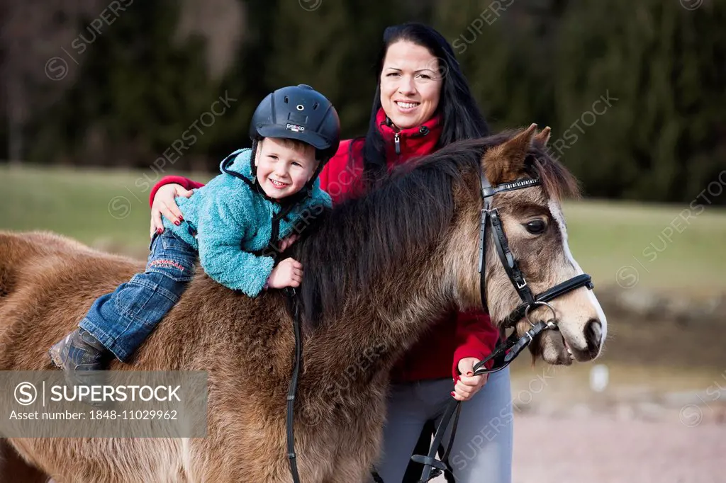 Young child wearing a riding helmet sitting bareback on a pony, dun, with a bridle, with a riding instructor, Tyrol, Austria