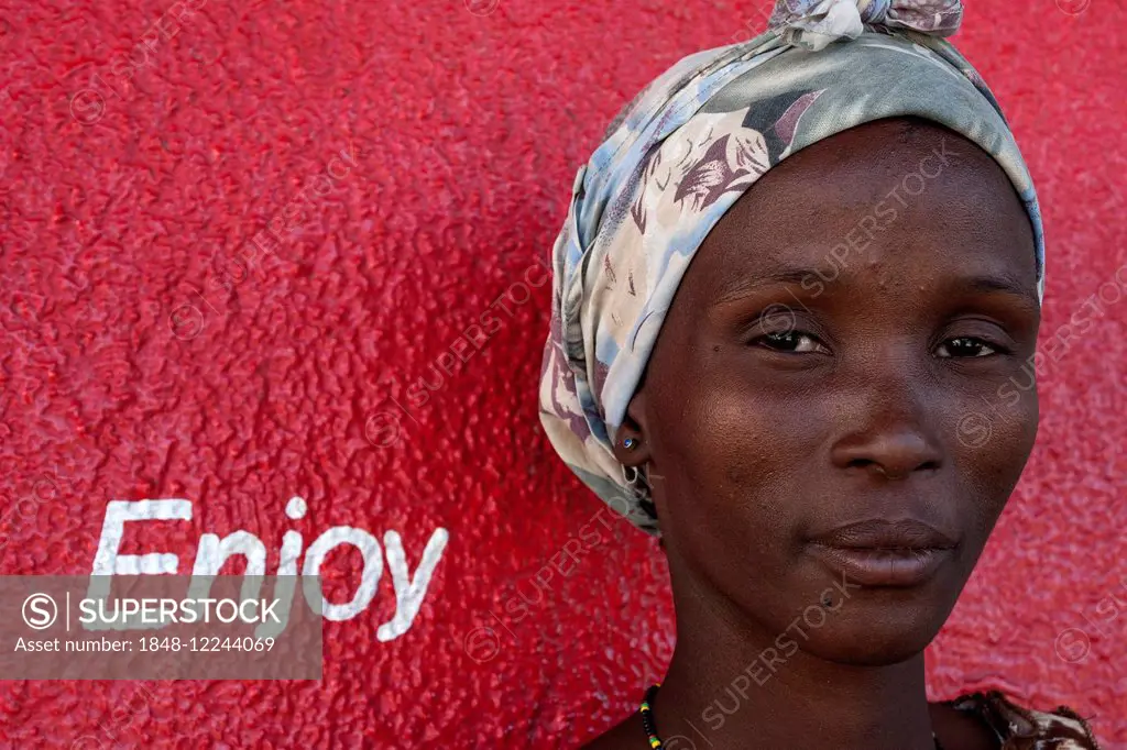 Local woman with a headscarf in front of a red wall with the lettering Enjoy, portrait, Outjo, Namibia