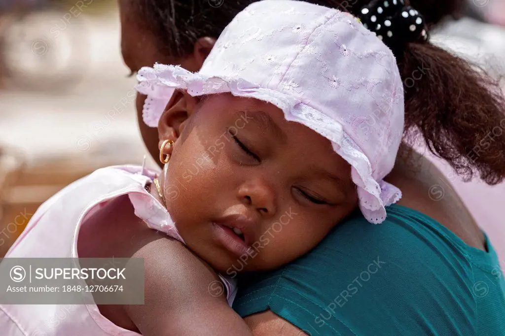 Little girl with white cap sleeping on her mother's shoulder, La Digue Island, Seychelles