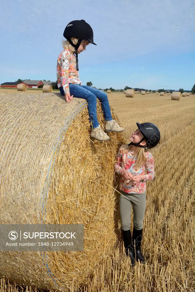 Two girls with riding hats, nine and four years old, at hay bale in field, Ystad, Scania, Sweden