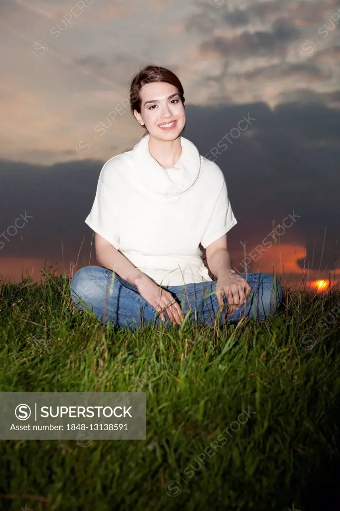Smiling young woman sitting in a meadow, sunset