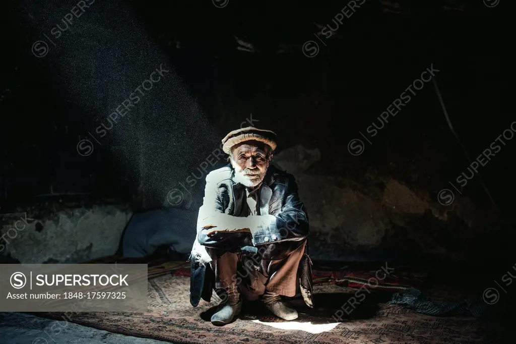 Man sitting on a carpet in his house, a beam of light falls through a skylight, Khas Dej, Wakhan corridor, Afghanistan, Asia