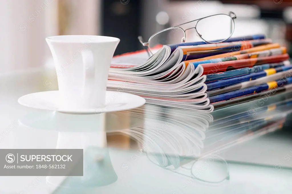 Still life with magazines, a coffee cup and a pair of glasses on a glass table