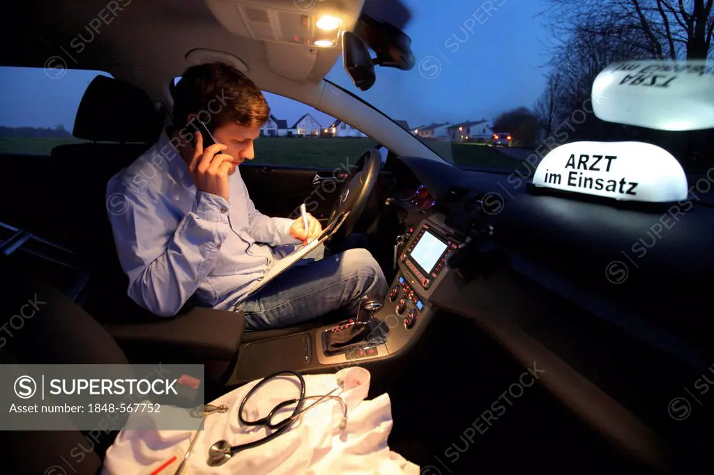 Young GP, general practitioner working in the country, making phone call and is taking notes in his car after an evening home visit, car displaying th...