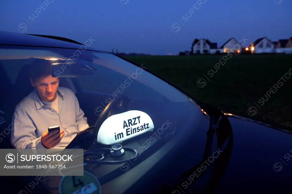 Young GP, general practitioner working in the country, making phone call in his car after an evening home visit, car displaying the sign Arzt im Einsa...