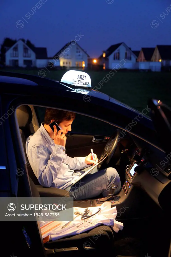Young GP, general practitioner working in the country, making phone call and taking notes in his car after an evening home visit, car displaying the s...