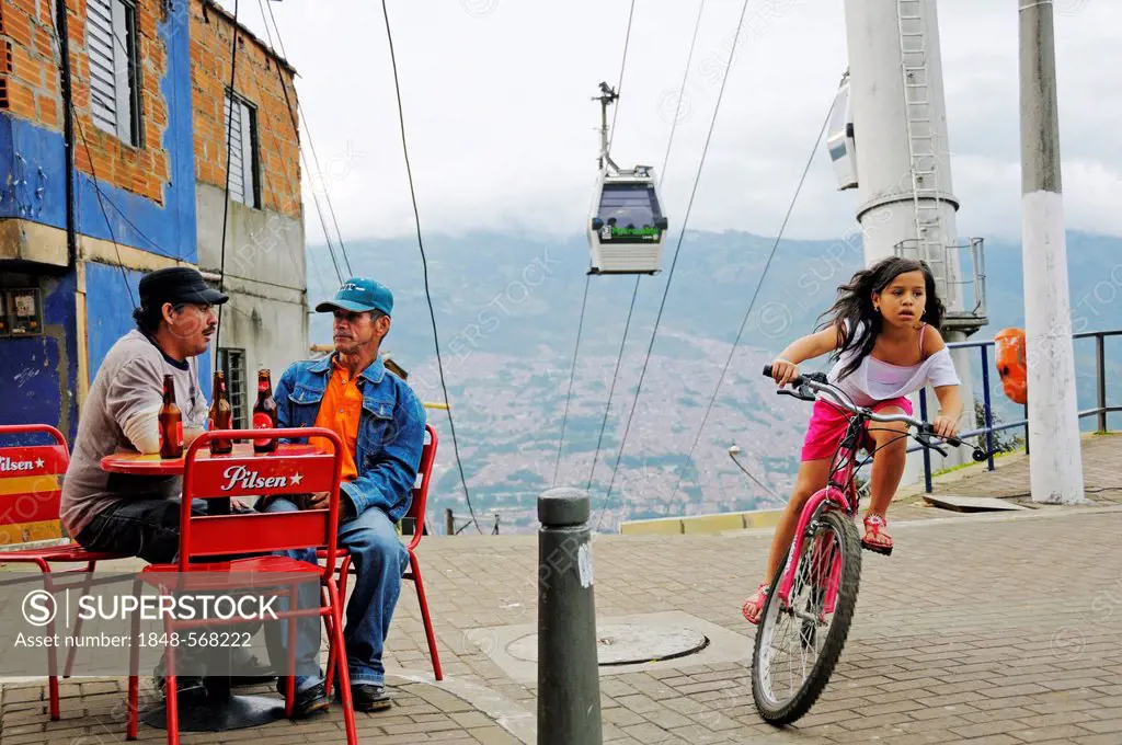 Men sitting in a street café, girl riding a bicycle, Metrocable cable car at back, slums, Comuna 13, Medellin, Colombia, South America, Latin America,...