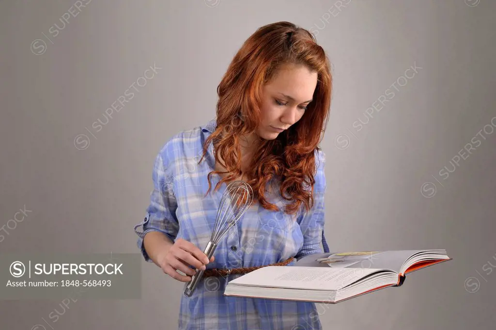 Young woman with red hair holding a whisk and a cook book in her hands