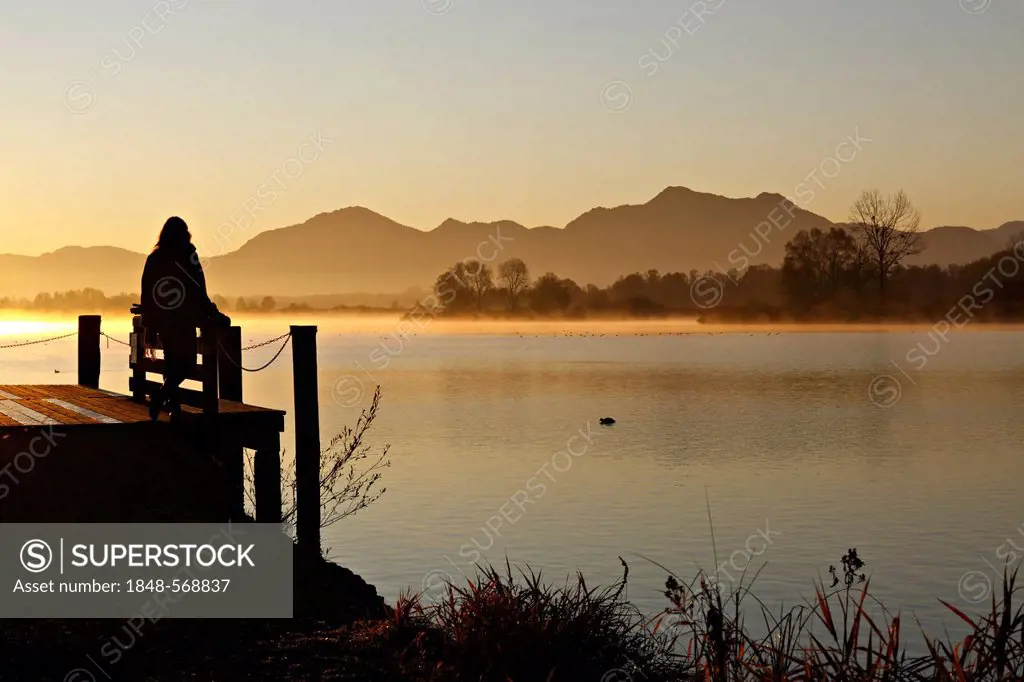 Silhouette of a woman sitting on a pier overlooking lake Chiemsee at sunrise, Chiemgau, Upper Bavaria, Germany, Europe