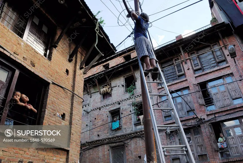 Young man standing on ladder that is leaning against power pole, Bhaktapur, Kathmandu Valley, Nepal, Asia