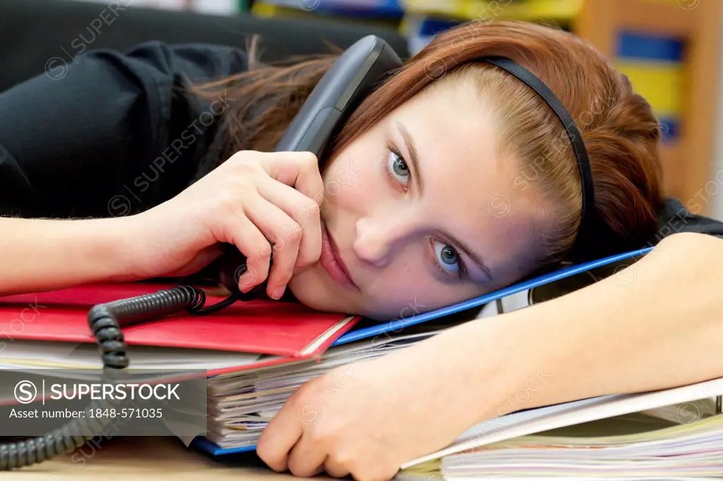 Exhausted office worker with telephone handset resting head on files and folders