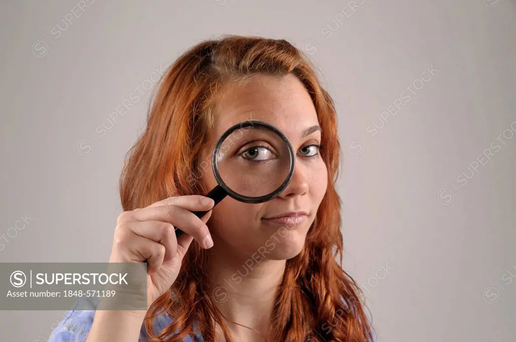 Young woman with red hair looking with one eye through a magnifying glass