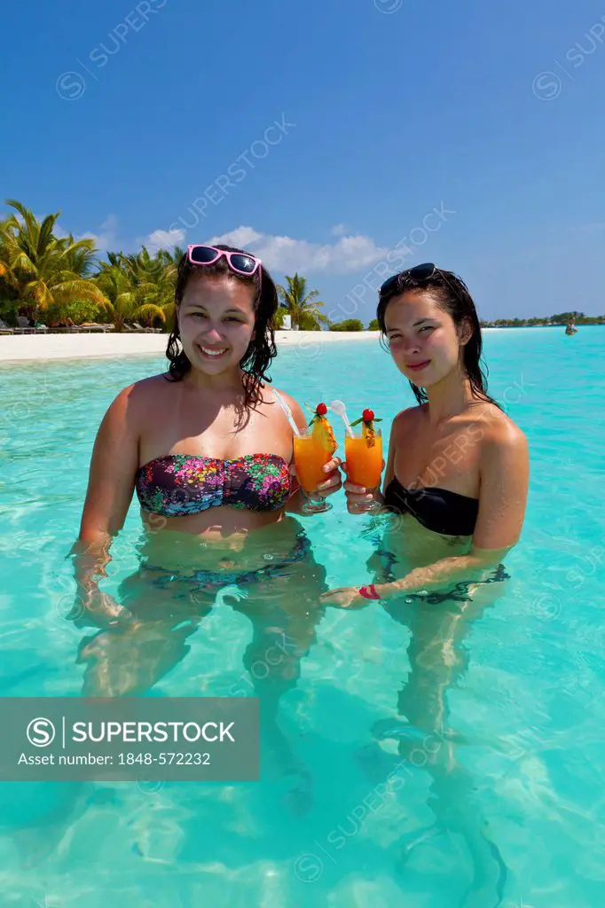 Two girls, about 14 and 18 years old, with sunglasses in their hair, drinking cocktails in a turquoise-coloured lagoon in the sea in front of an islan...