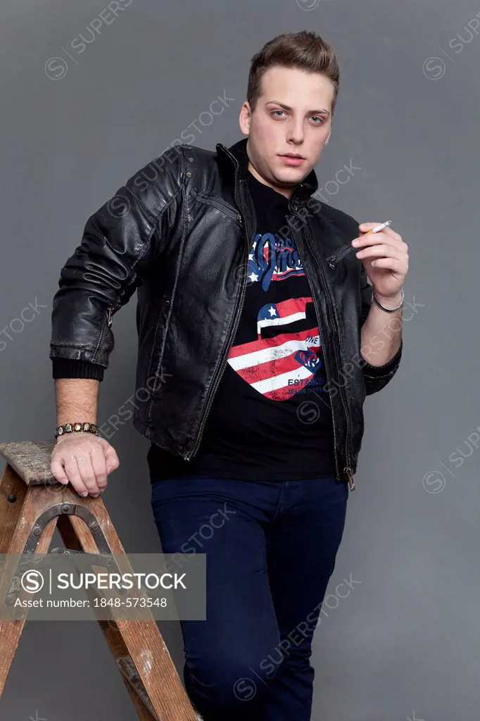 Young man in a leather jacket and jeans smoking a cigarette whilst leaning on an old wooden ladder