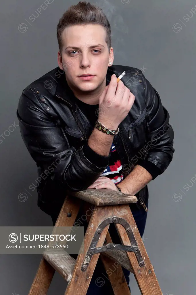 Young man in a leather jacket smoking a cigarette whilst resting on an old wooden ladder