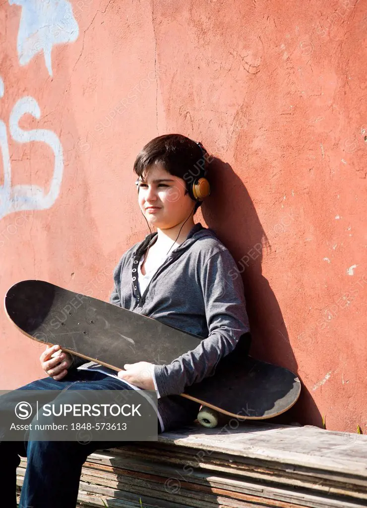 Boy wearing headphones and holding a skateboard sitting in front of a wall