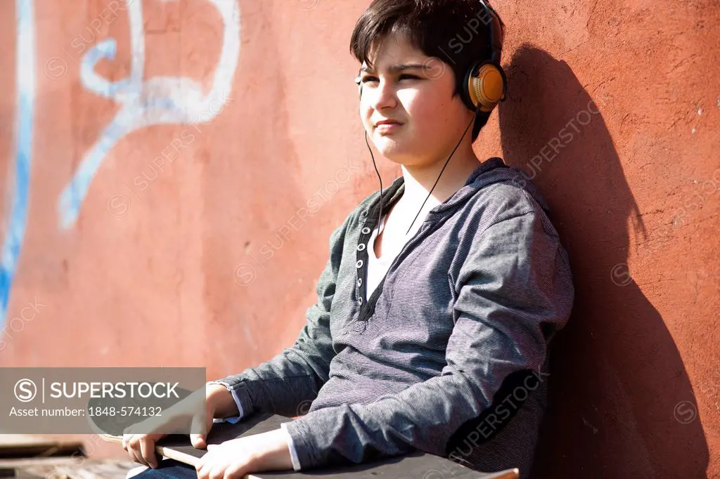 Boy wearing headphones and holding a skateboard sitting in front of a wall