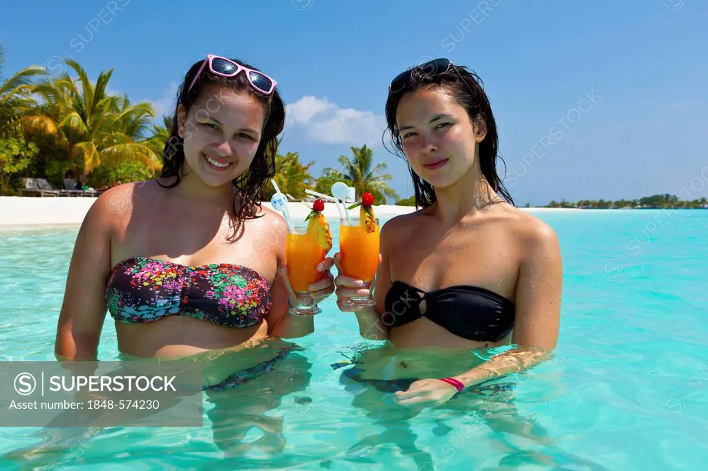 Two girls, about 14 and 18 years old, with sunglasses in their hair, drinking cocktails in a turquoise-coloured lagoon in the sea in front of an islan...