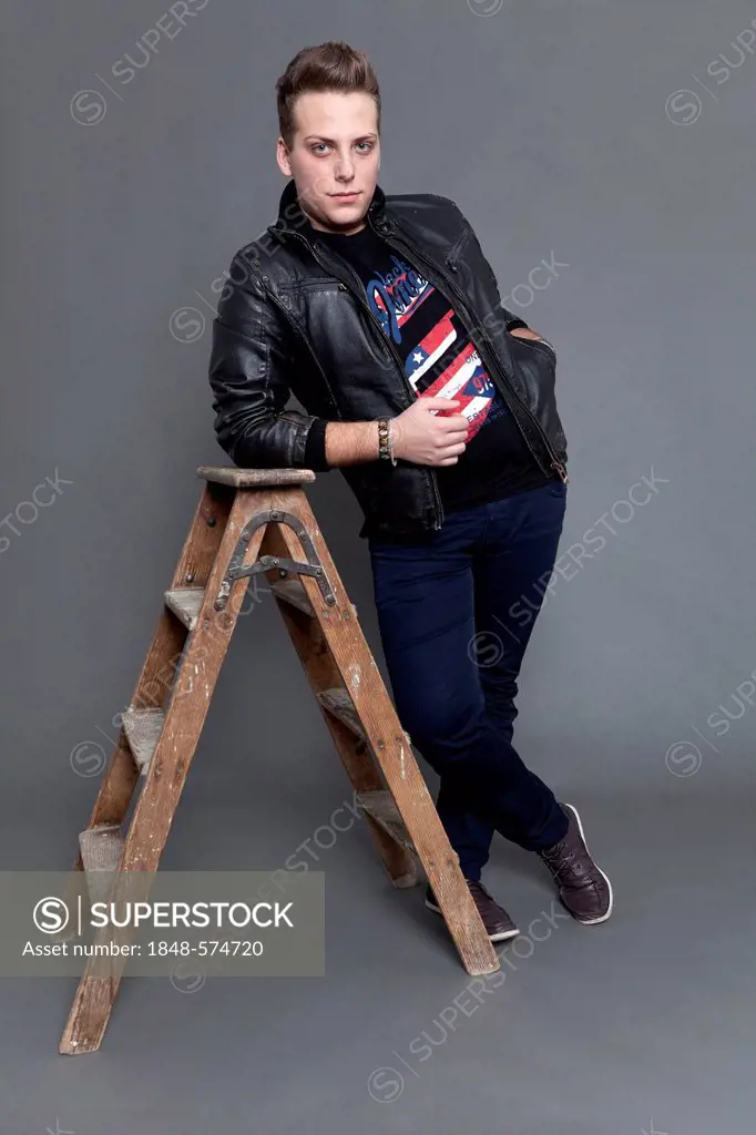 Young man wearing a leather jacket and jeans, leaning against an old wooden ladder