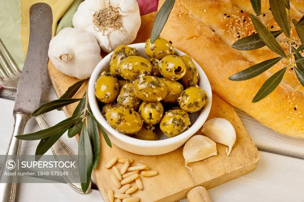 A bowl of marinated olives on a wodden board with bread, garlic and cutlery