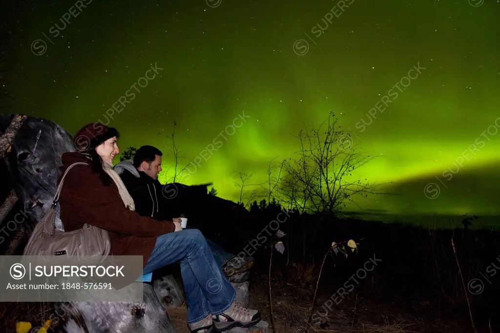 Couple, woman and man, sitting in a wooden chair, watching swirling Northern lights, Polar Aurorae, Aurora Borealis, green, near Whitehorse, Yukon Ter...