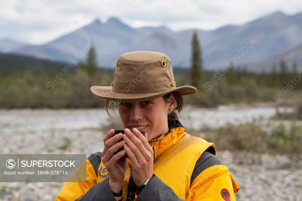 Young woman drinking, enjoying a cup of tea, Wind River, Peel Watershed, Northern Mackenzie Mountains behind, Yukon Territory, Canada