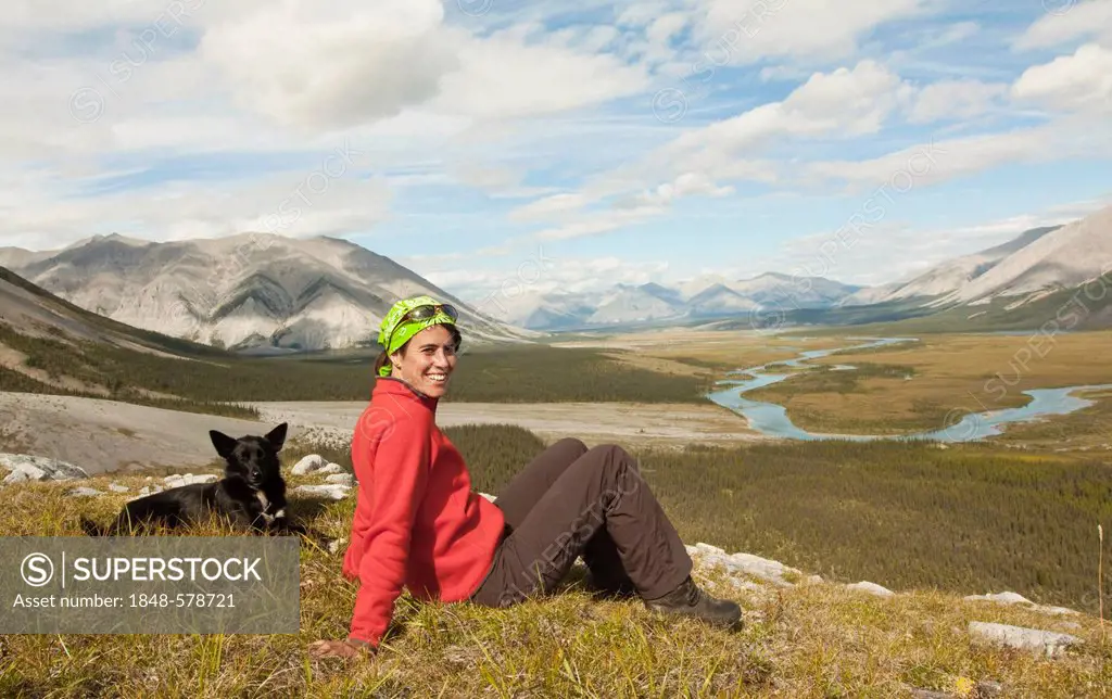 Young woman sitting, resting, enjoying the view, panorama, Alaskan Husky, sled dog beside her, Wind River valley and Mackenzie Mountains behind, Peel ...