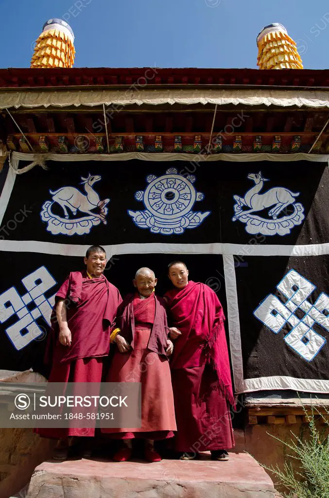 Three Tibetan Buddhist nuns, a younger, a middle-aged and a 70-year-old woman in red monks' robes in front of the Tibetan Buddhist symbols at the mona...