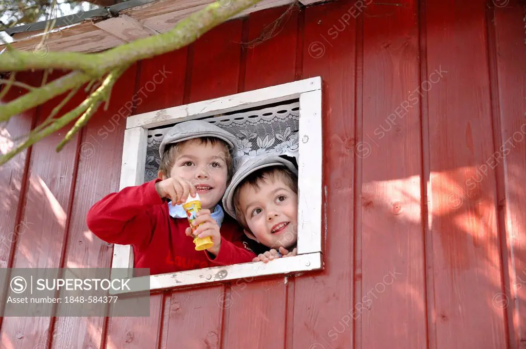 Twin boys, 4, wearing flat caps and blowing soap bubbles from the window of a tree house