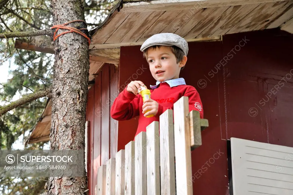 Boy, 4, wearing a flat cap and blowing soap bubbles in front of a tree house