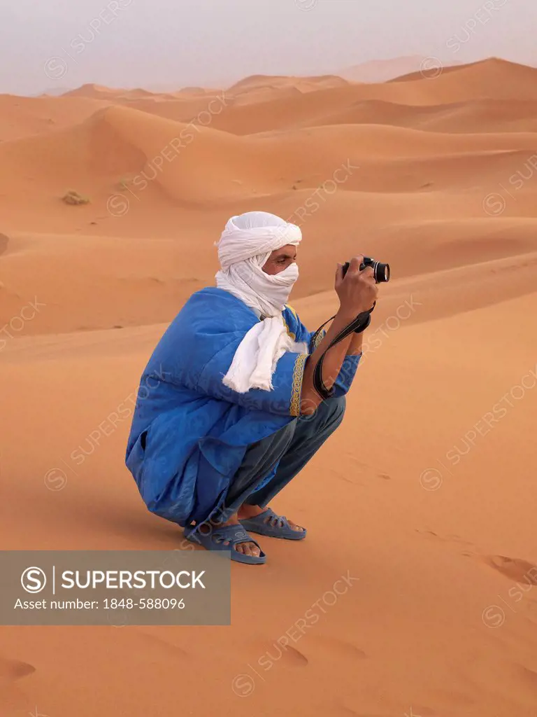 Tuareg man dressed in a blue robe and a white turban taking a photograph in the sand dunes of the Erg Chebbi Desert, near Merzouga, Morocco, North Afr...