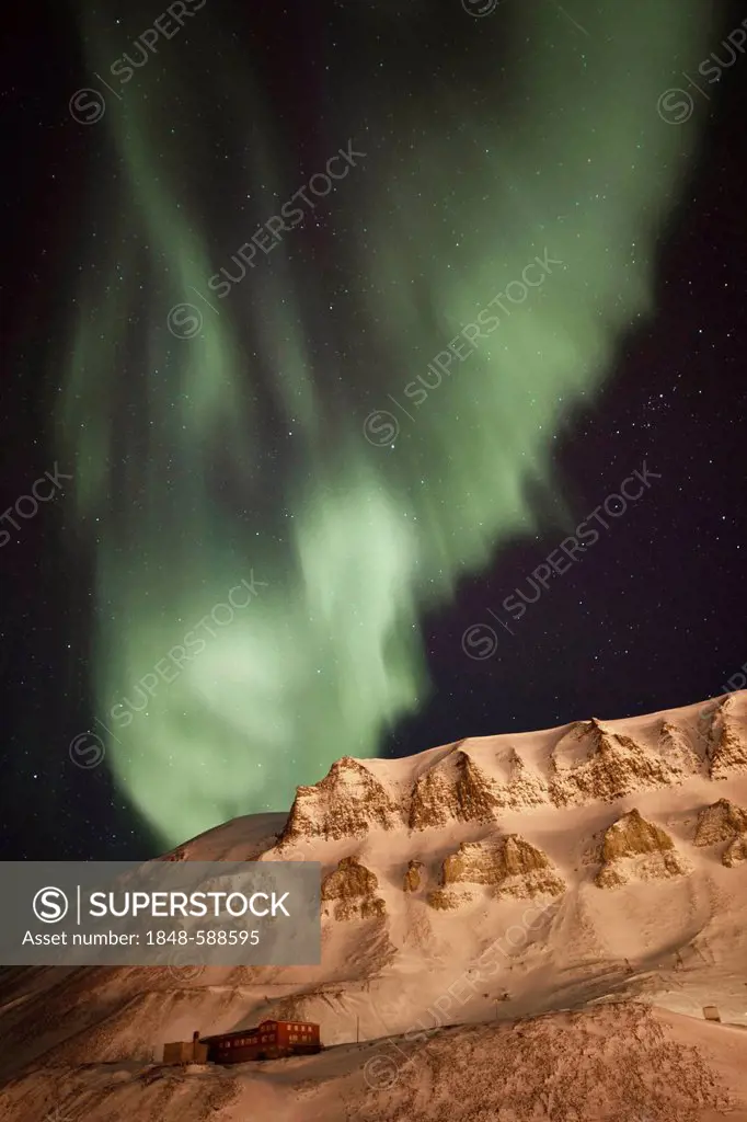 Strong green Northern Lights, Aurora Borealis, above a historic building lit by the lights of the town of Longyearbyen, Longyearbreen Glacier at back,...