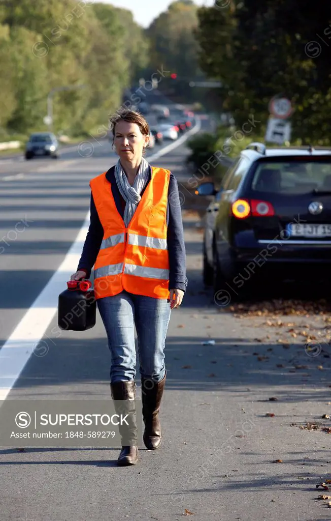 Car breakdown, female driver has stopped on the hard shoulder of a country road, wearing a reflective vest carrying an empty jerry can, on her way to ...