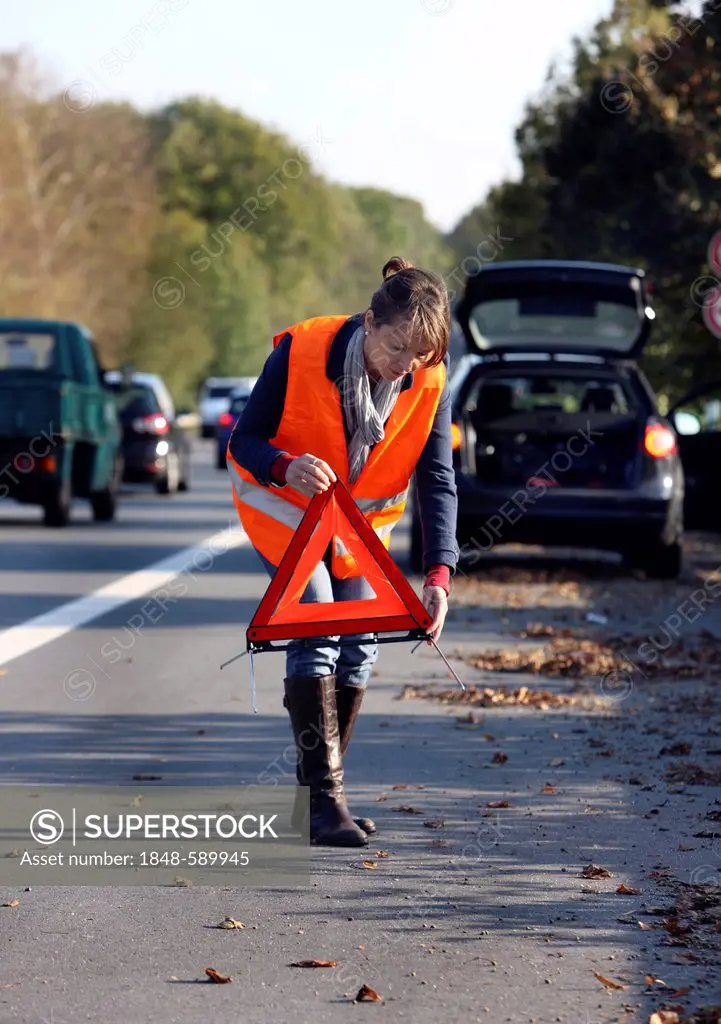 Car breakdown, female driver has stopped on the hard shoulder of a country road, wearing a reflective vest, putting up a warning triangle