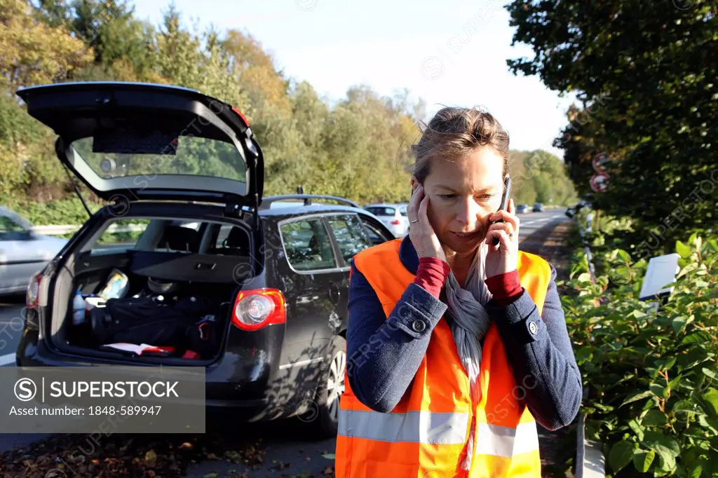 Car breakdown, female driver has stopped on the hard shoulder of a country road, wearing a reflective vest, makes call for help on her mobile phone