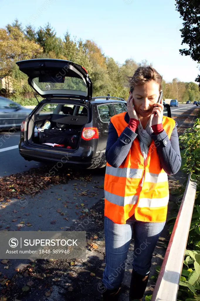 Car breakdown, female driver has stopped on the hard shoulder of a country road, wearing a reflective vest, makes call for help on her mobile phone