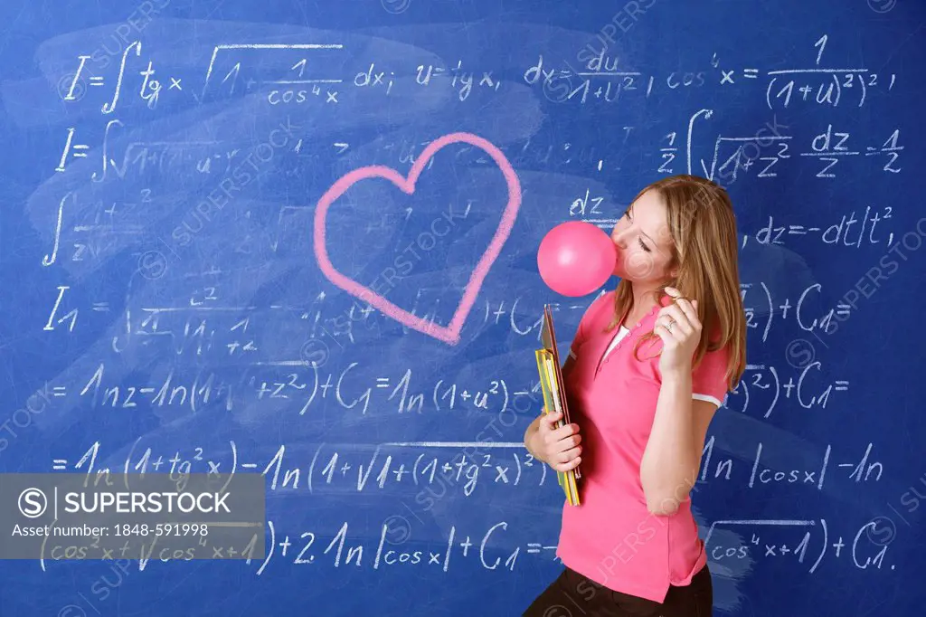 Schoolgirl daydreaming while blowing a bubble with bubble gum in front of a school blackboard with a drawn heart and arithmetic problems