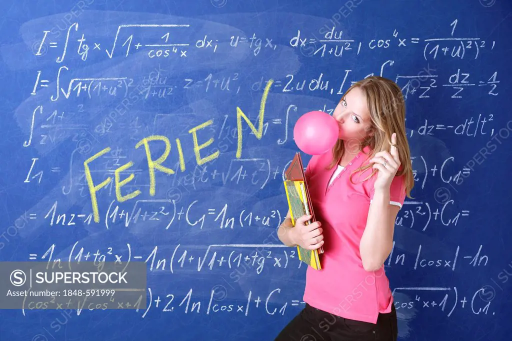 Schoolgirl blowing a bubble with bubble gum in front of a school blackboard with the word Ferien, German for holidays, and arithmetic problems