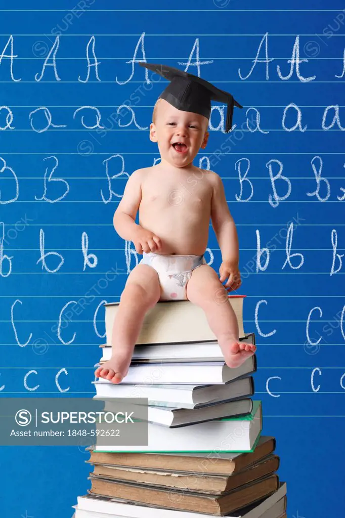 Laughing infant wearing a graduation cap sitting on a pile of books in front of a school blackboard with the ABC