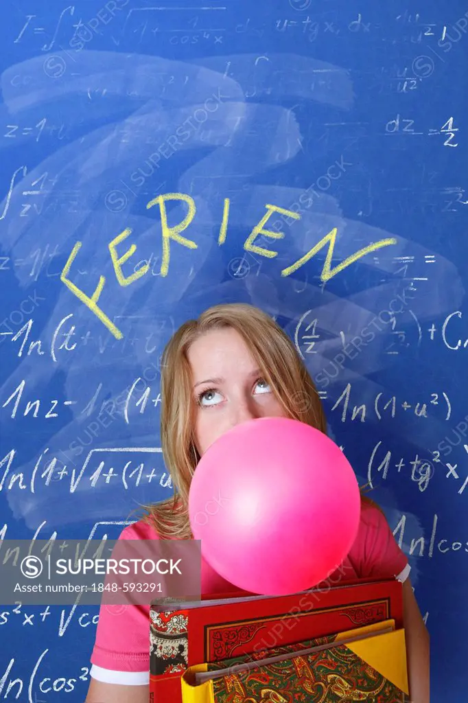 Schoolgirl blowing a bubble with bubble gum in front of a school blackboard with the word Ferien, German for holidays, and arithmetic problems