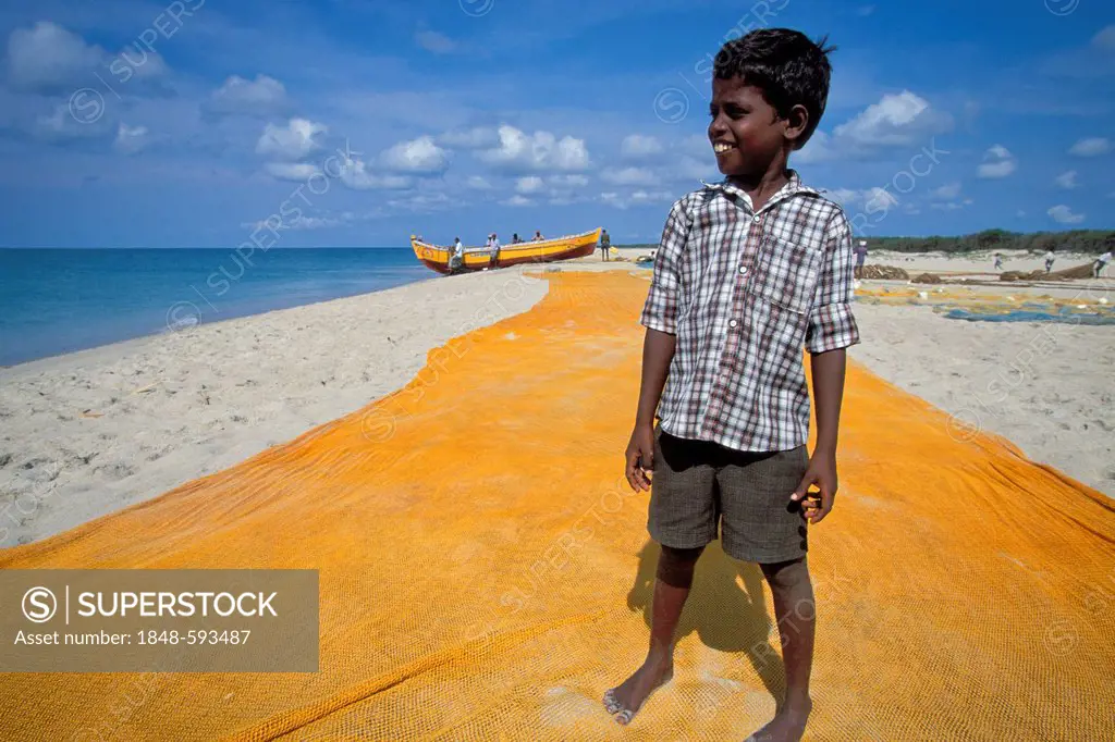 Boy, son of a fisherman, standing on a yellow fishing net in front of a fishing boat, Adam's Bridge, near Rameshwaram or Ramesvaram, Tamil Nadu, South...