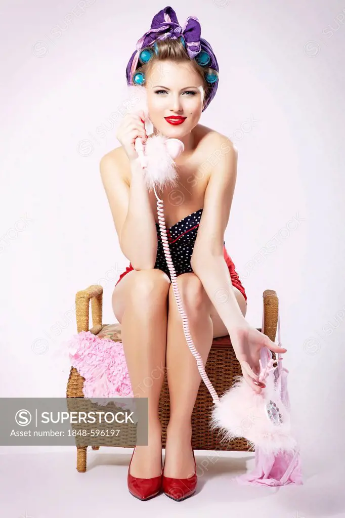 Young woman with hair curlers and hot pants, sitting on a laundry basket and talking on the phone, pin-up