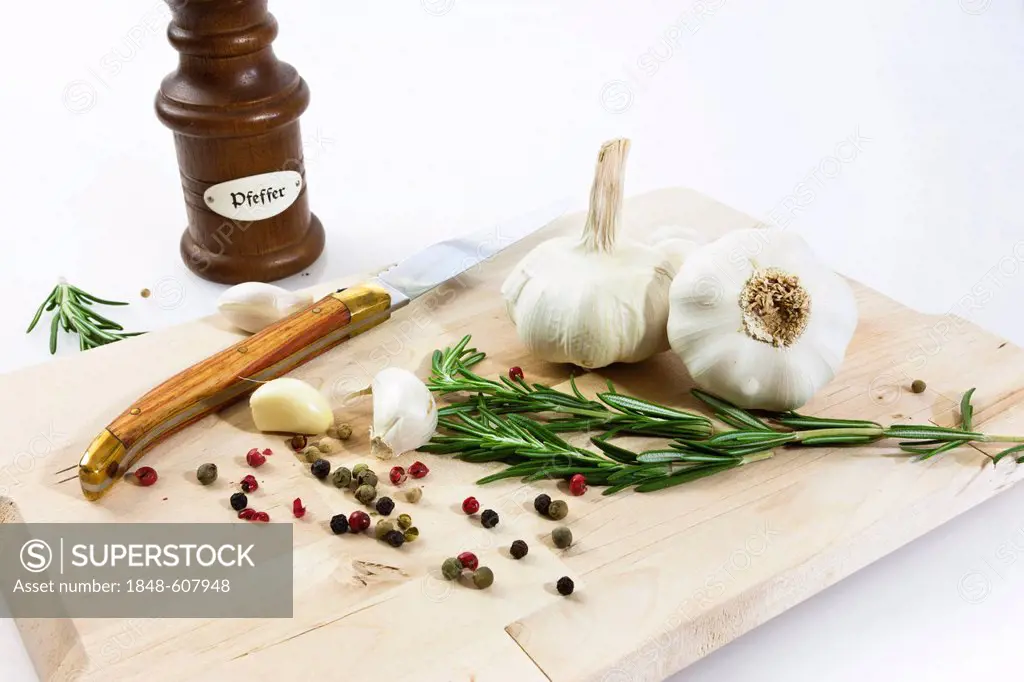 Garlic (Allium sativum) on a wooden board with knife, pepper mill, rosemary and black and red pepper