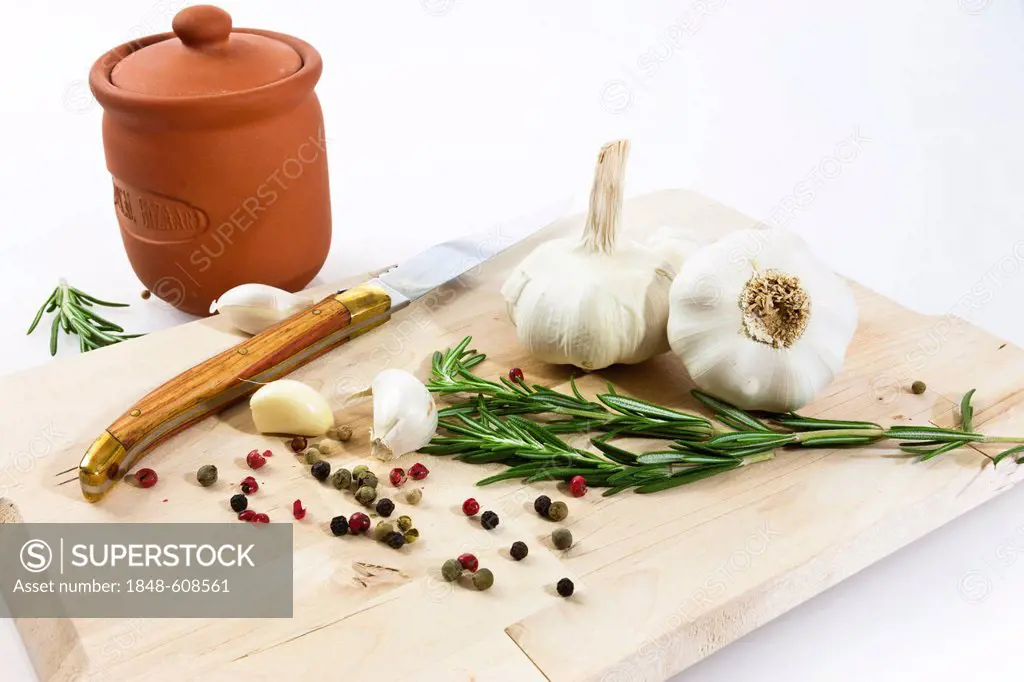 Garlic (Allium sativum) on a wooden board with knife, clay pot, rosemary and black and red pepper