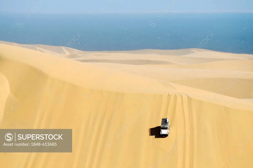 Landrover Defender off-road vehicle driving down a huge dune, Atlantic Ocean at the back, Namib Naukluft National Park, part of the Namibian Skeleton ...