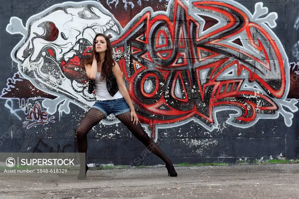 Young woman in a white top, denim hot pants, black stockings and high heels posing in front of a graffiti wall