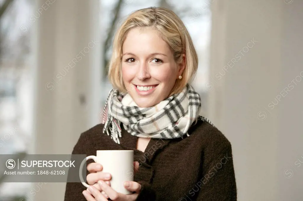 Smiling young blonde woman in a dark sweater and wool scarf holding a mug