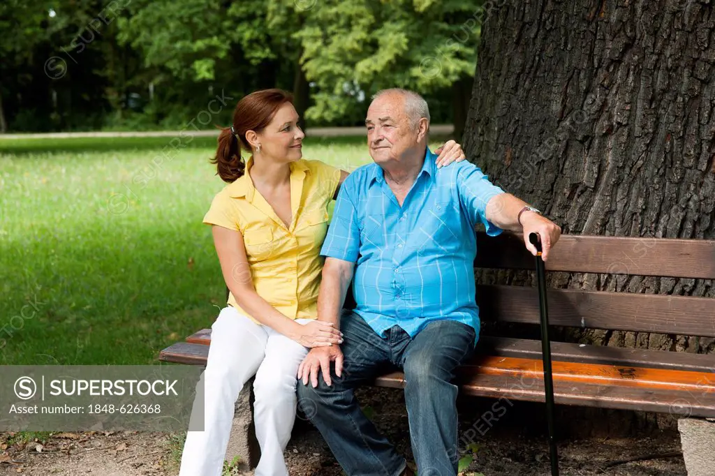 A woman and an elderly man with a walking cane sitting on a bench under a tree