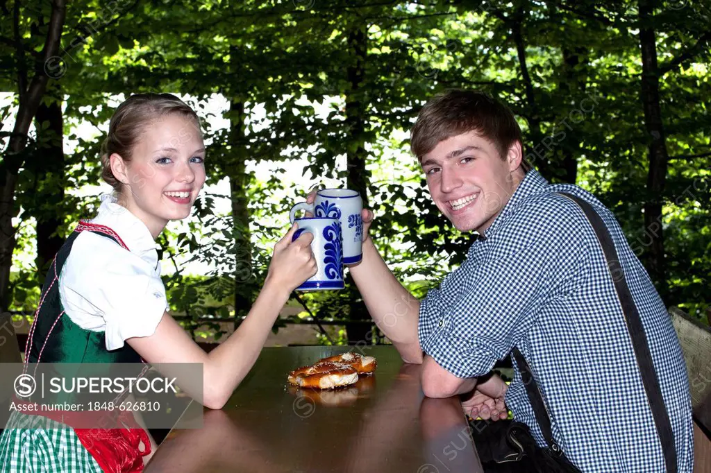 Young couple in dirndl and lederhosen clinking their beer steins, beer garden, at Pettstatt, Upper Franconia, Bavaria, Germany, Europe